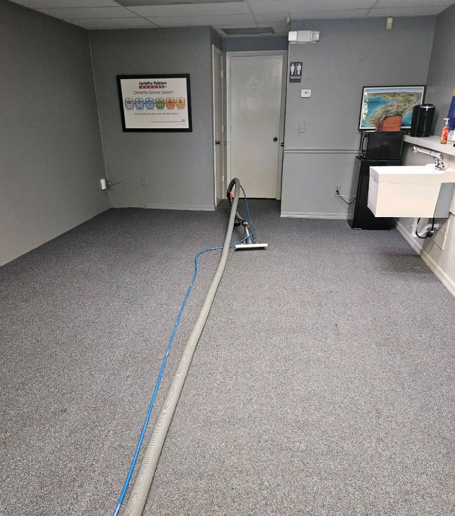 Empty office room with gray carpet, a cord on the floor, and wall art near a closed door.