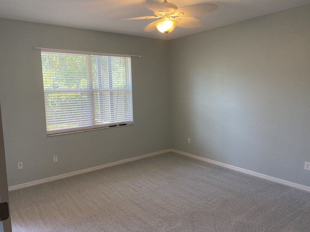 Empty gray bedroom with carpet, window blinds, and a ceiling fan lighting the room