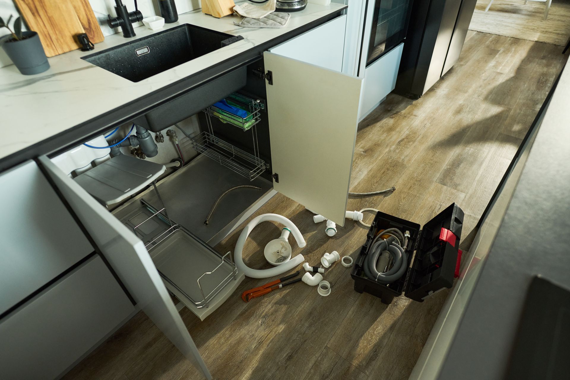 Under-sink plumbing cabinet with exposed pipes, cabinet doors open, and a power tool on the floor