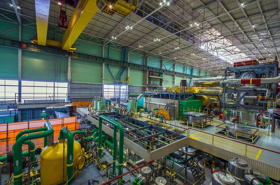Large industrial facility with complex machinery, piping, and overhead crane. Green, yellow, and silver equipment.