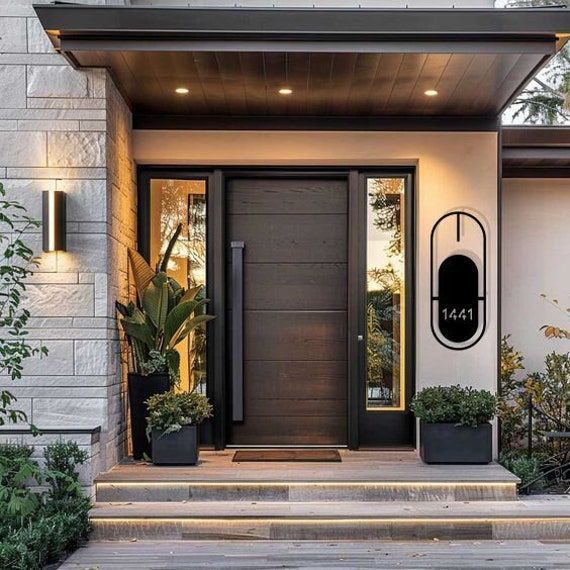 Modern home entrance with dark wooden door, glass sidelights, and address plaque. Lit steps, plants.