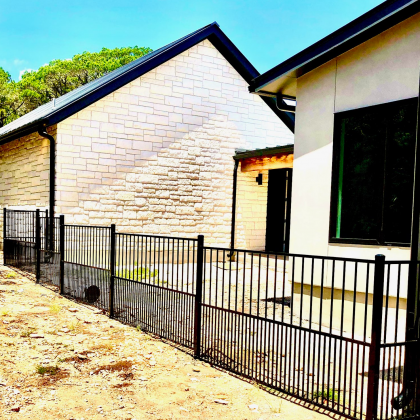 Black metal fence in front of two buildings with light-colored stone and stucco walls, under blue sky.