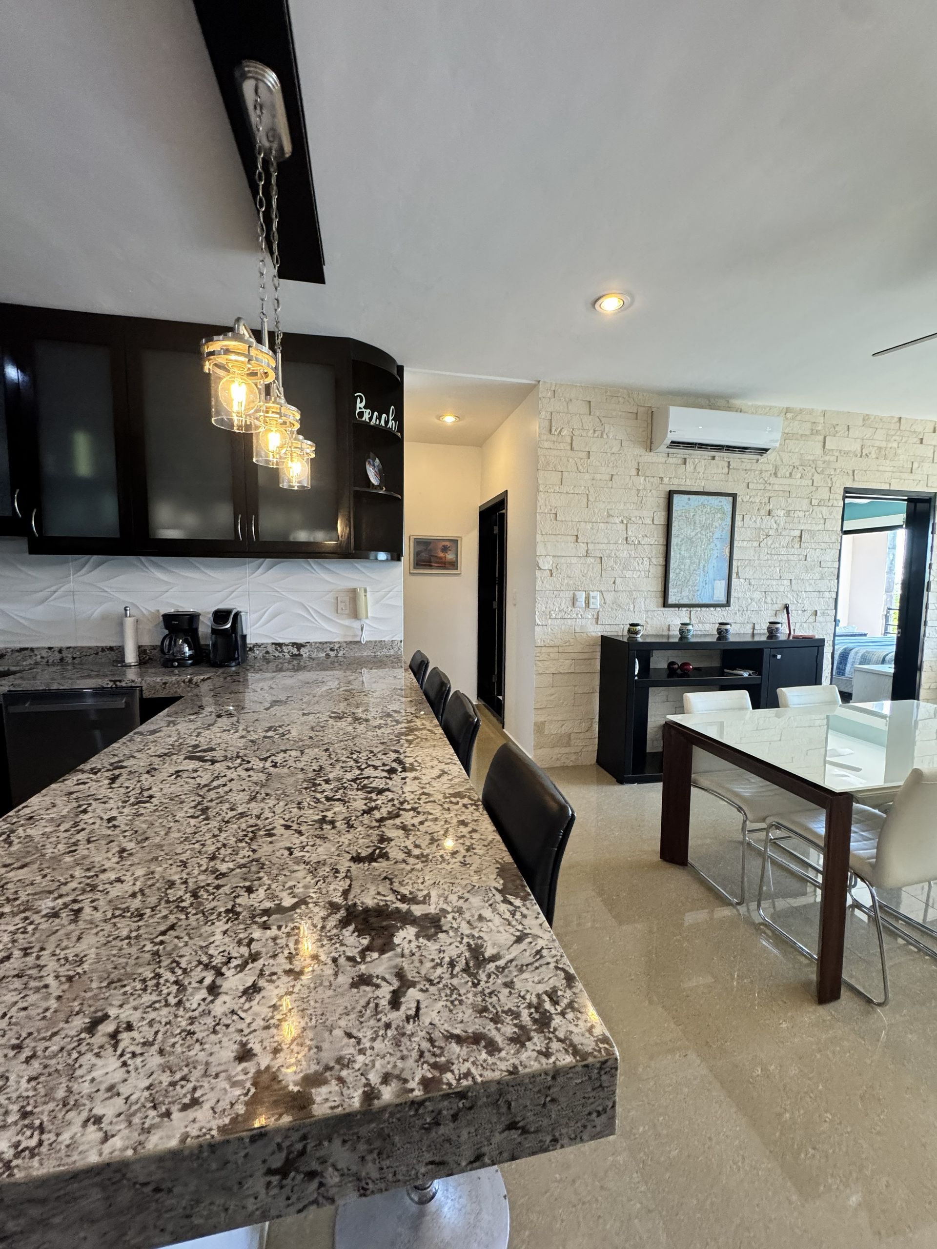 Cocina moderna con isla de granito, armarios negros, lámparas colgantes y mesa de comedor en un interior de planta abierta.