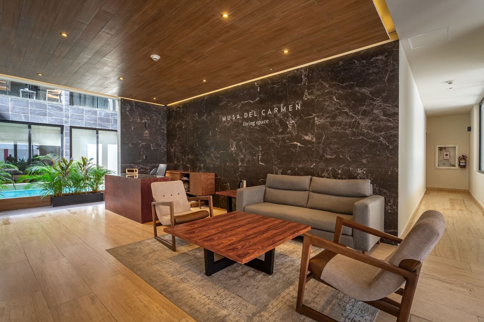 Vestíbulo de hotel moderno con sofá gris, sillas de madera, mesa de centro y pared de mármol detrás de la zona de recepción.