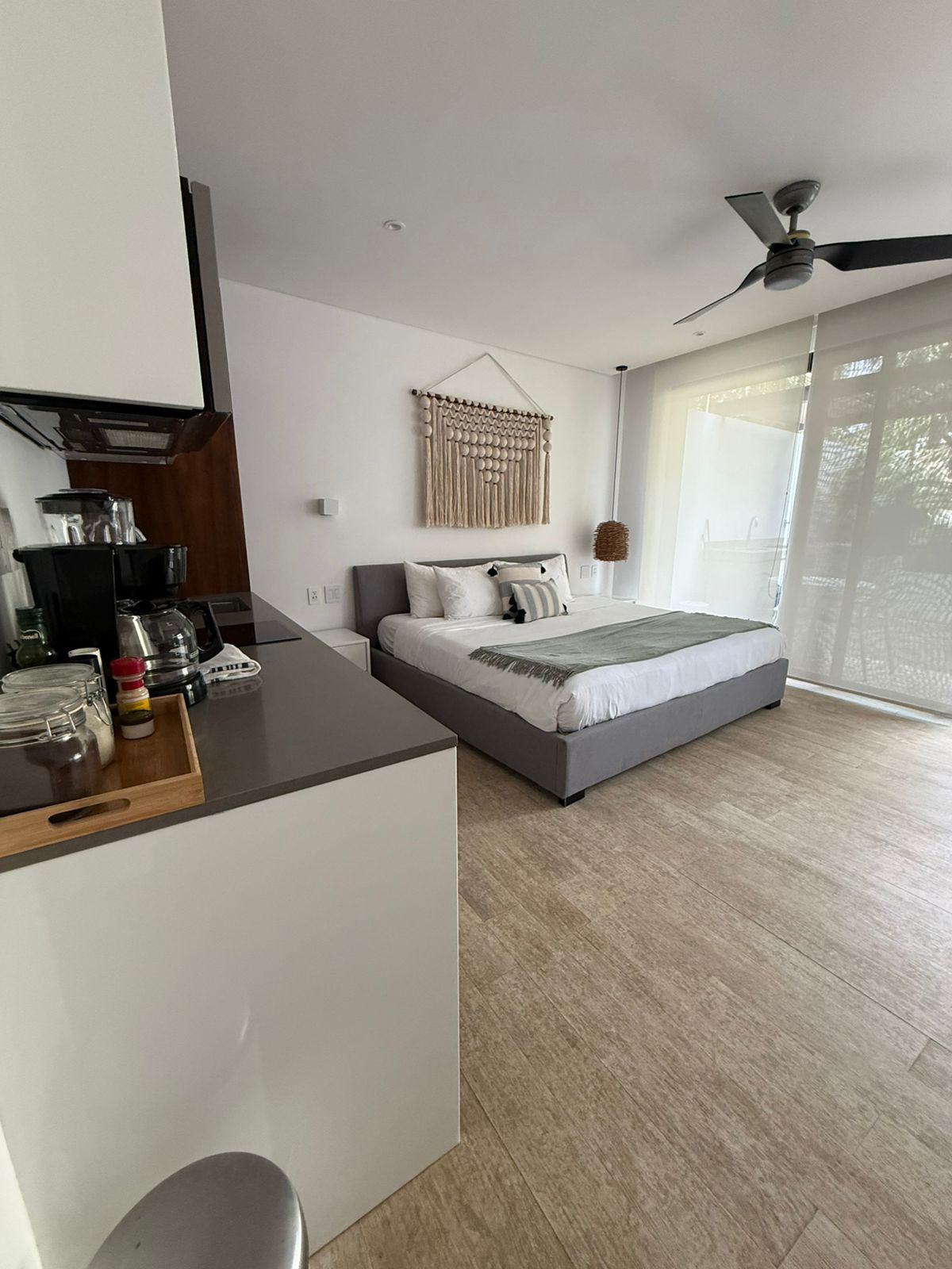 Habitación luminosa con cama gris, decoración de pared tejida, ventilador de techo y puertas corredizas de vidrio que dan al exterior.
