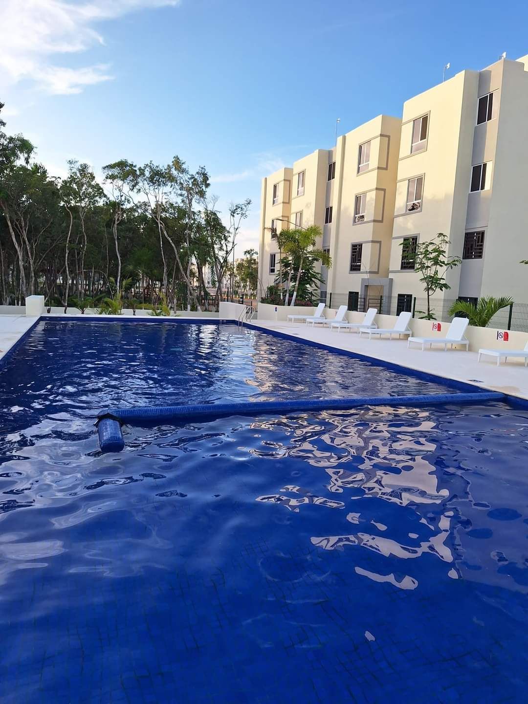 Piscina al aire libre junto a un edificio de apartamentos beige bajo un cielo azul claro.