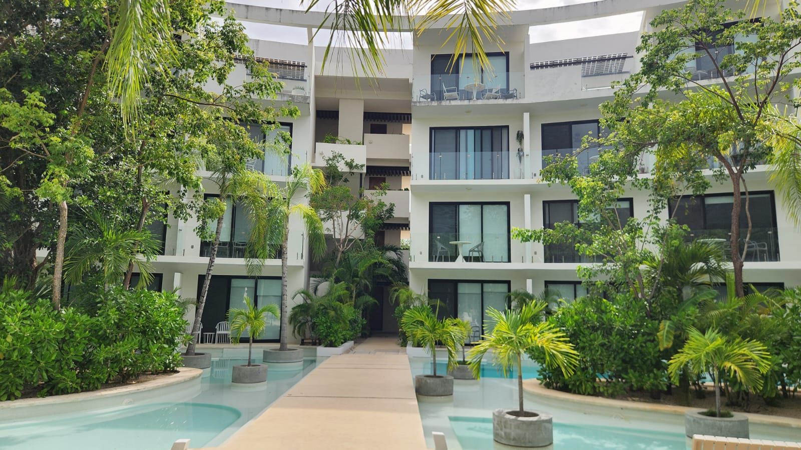 Patio interior de apartamentos modernos con piscinas, palmeras y balcones alrededor de un paseo central.