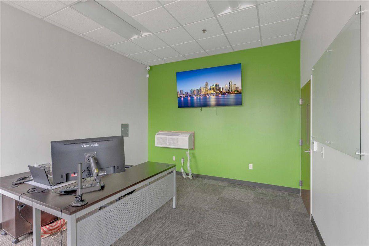 Office with desk, computer, TV displaying cityscape, green accent wall, whiteboard, and gray carpet.