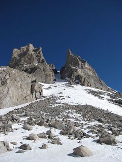 A steep, snow-covered mountain slope leading up to two sharp, rocky peaks against a clear, deep blue sky.