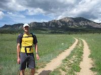 A person wearing a yellow shirt, shorts, and a cap smiles while hiking on a dirt trail toward mountains under a cloudy sky.