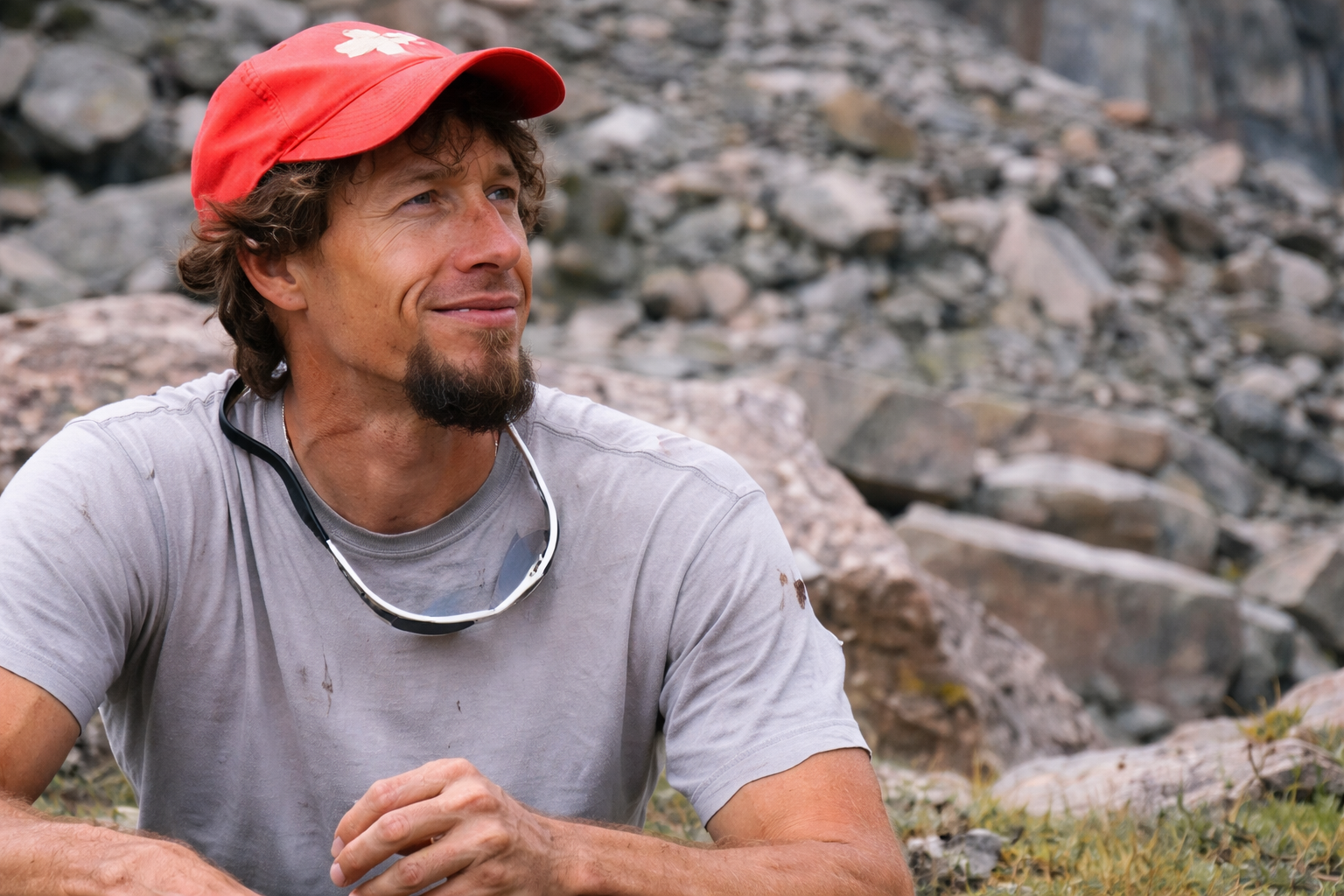 A person with a goatee wearing a red cap and grey t-shirt sits outdoors against a rocky background, looking to the side.