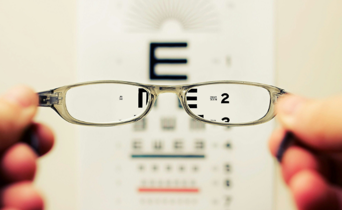 Person holding glasses in front of an eye chart. The glasses improve clarity of the chart's letters.