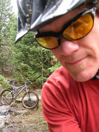 Cyclist in red shirt and helmet close to camera, with bike on a rocky forest trail in the background