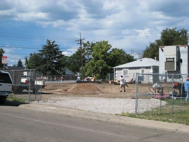 Construction site with gravel lot, chain-link fence, workers, and a white tent under a blue sky