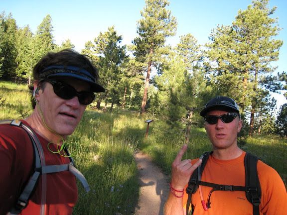 Two hikers on a forest trail, wearing sunglasses and backpacks, with trees and sunlight in the background