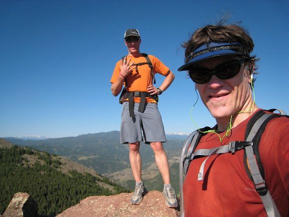 Two hikers on a rocky summit, wearing orange packs with mountains in the background.