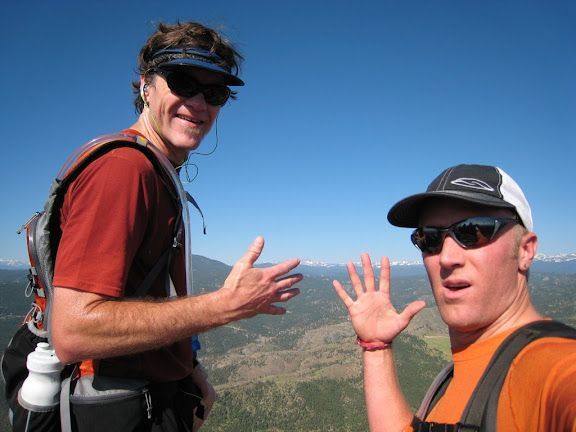 Two hikers in sunglasses pose on a mountain trail, smiling with a scenic valley behind them.