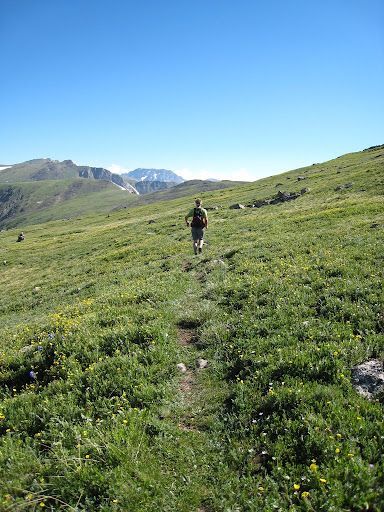 Hiker on a narrow trail through green alpine hills under a clear blue sky