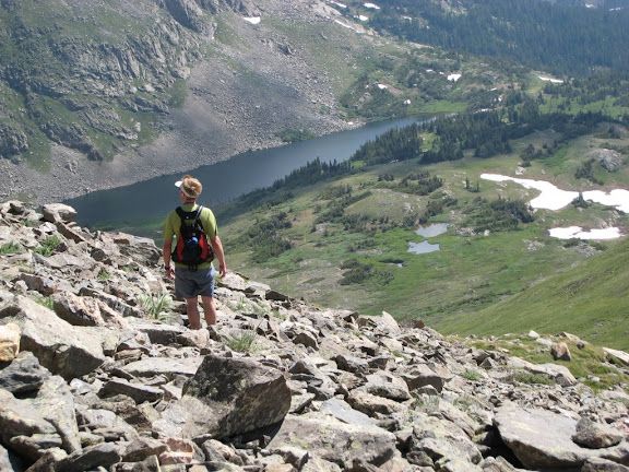 Hiker on a rocky mountain slope overlooking a valley lake with patches of snow