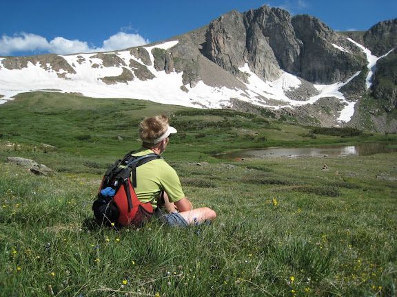 Hiker with red backpack sitting in a green alpine meadow below snowy mountain peaks