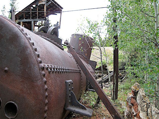 Rusty industrial machinery outdoors amid trees and a wooden shelter