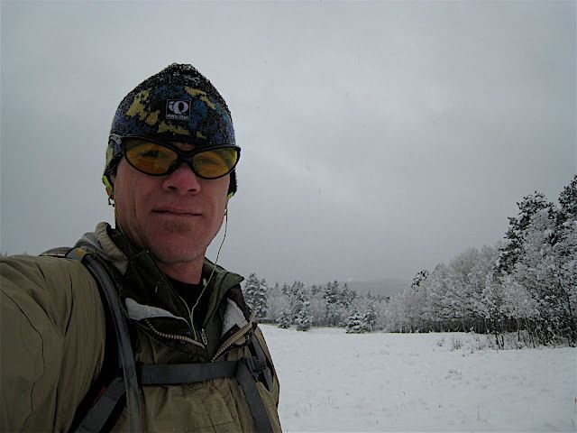 Person in a beanie and sunglasses taking a selfie in a snowy landscape with trees and overcast sky