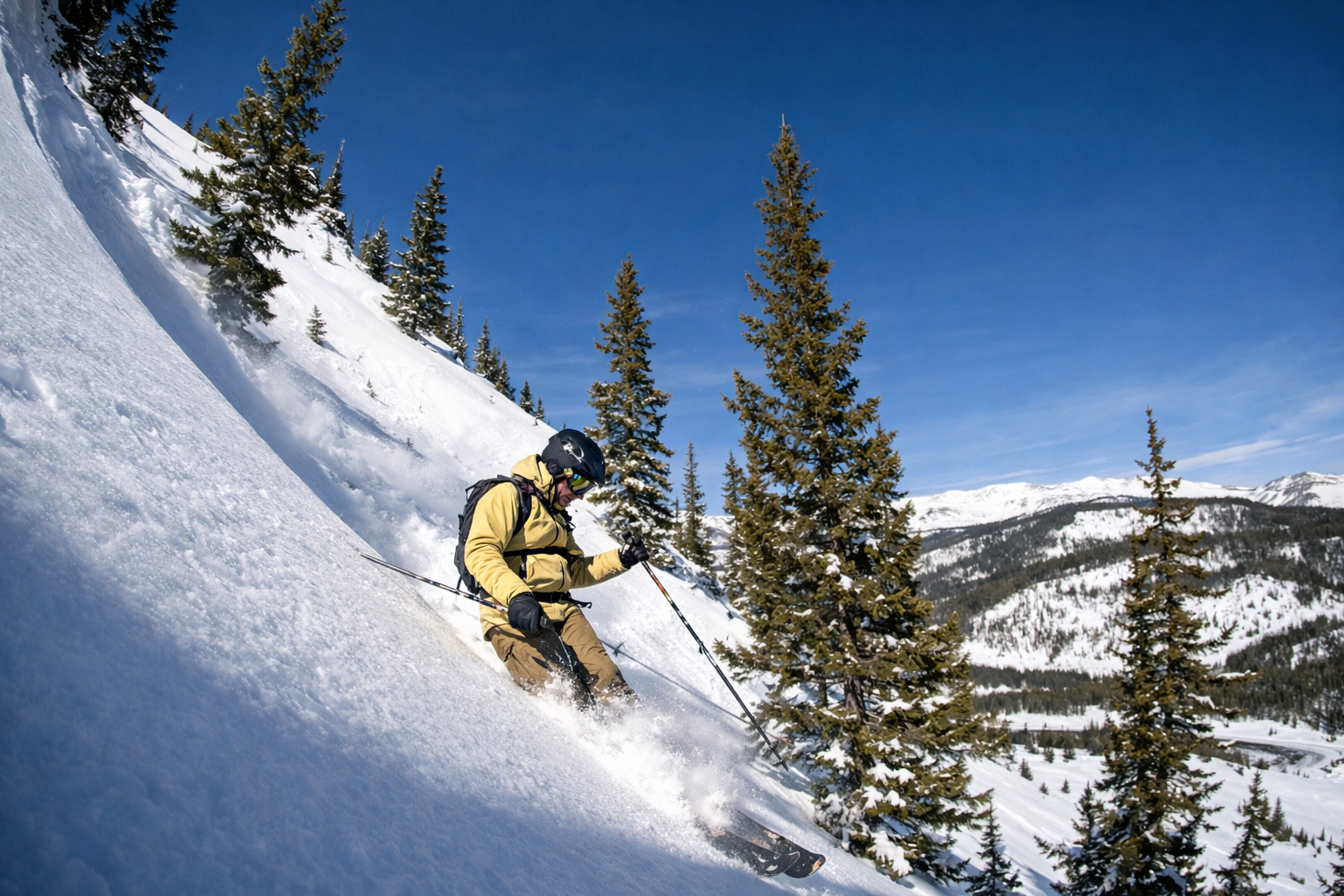 A person in a yellow jacket skis down a steep, snow-covered mountain slope surrounded by pine trees under a clear blue sky.
