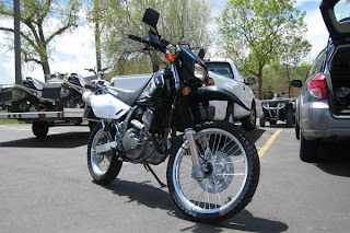 Black and white dirt bike parked in a lot beside cars, with trees in the background.
