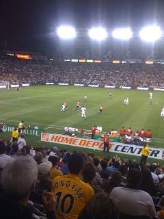 Soccer match at a crowded stadium under bright lights, with a fan in a yellow jersey in the foreground.