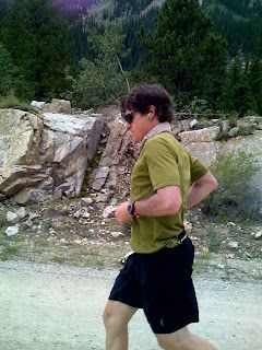Person jogging on a rocky trail beside trees and a mountain backdrop.