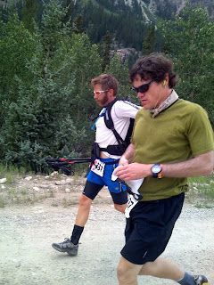 Two runners in athletic gear running on a gravel trail beside a forest.