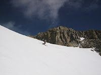 Snow-covered mountain slope beneath rocky cliffs and a cloudy blue sky