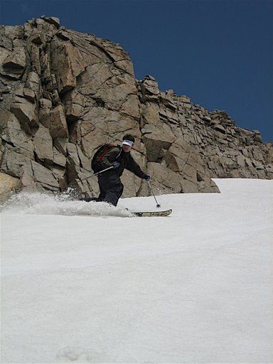 Skier carving down a snowy slope beside a rocky cliff under a clear blue sky