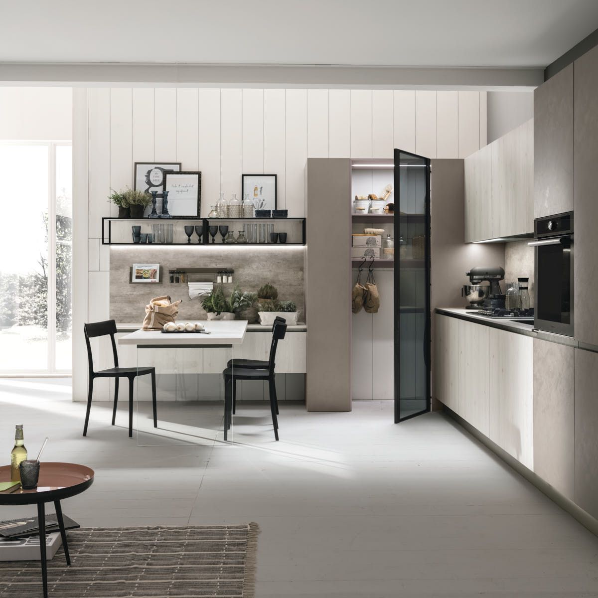Cucina moderna con mobili in legno chiaro, tavolo con sedie e dispensa con anta a vetri.