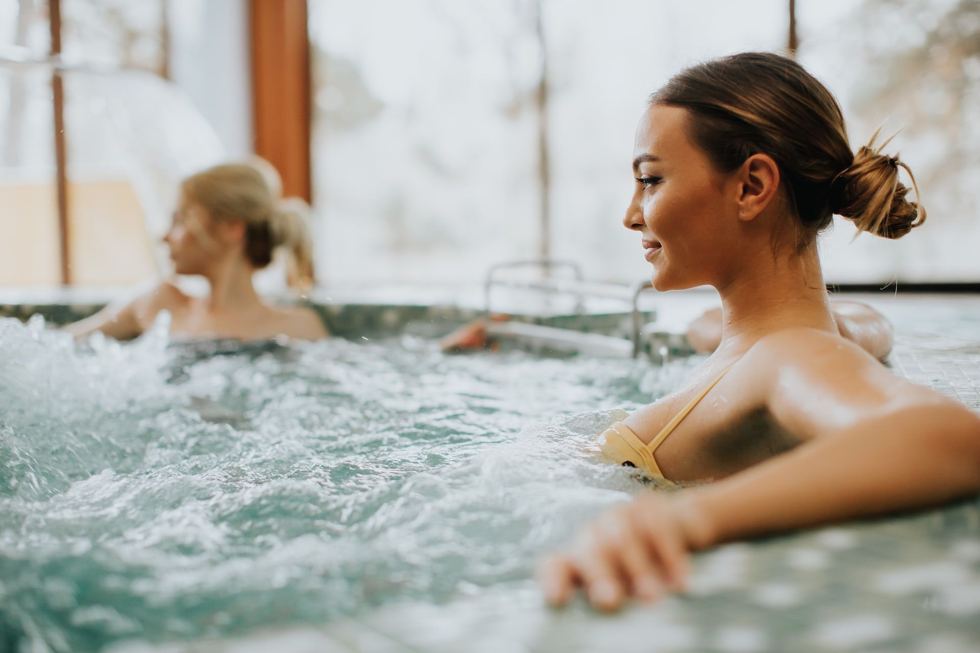 Two women relax in a bubbling hot tub; one smiles, water splashes around them.