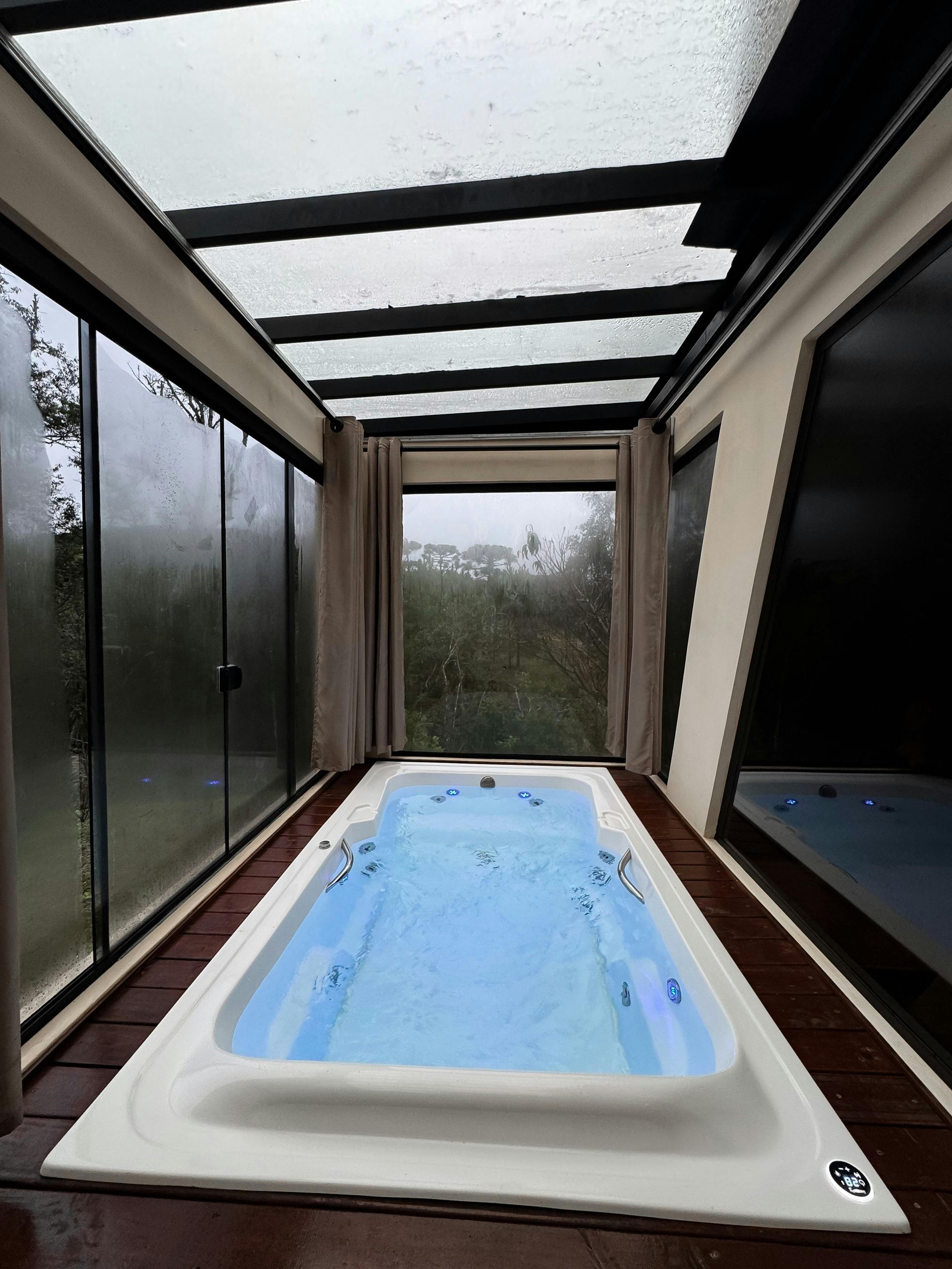 Indoor rectangular spa with blue water, enclosed by windows and a glass ceiling, overlooking greenery.