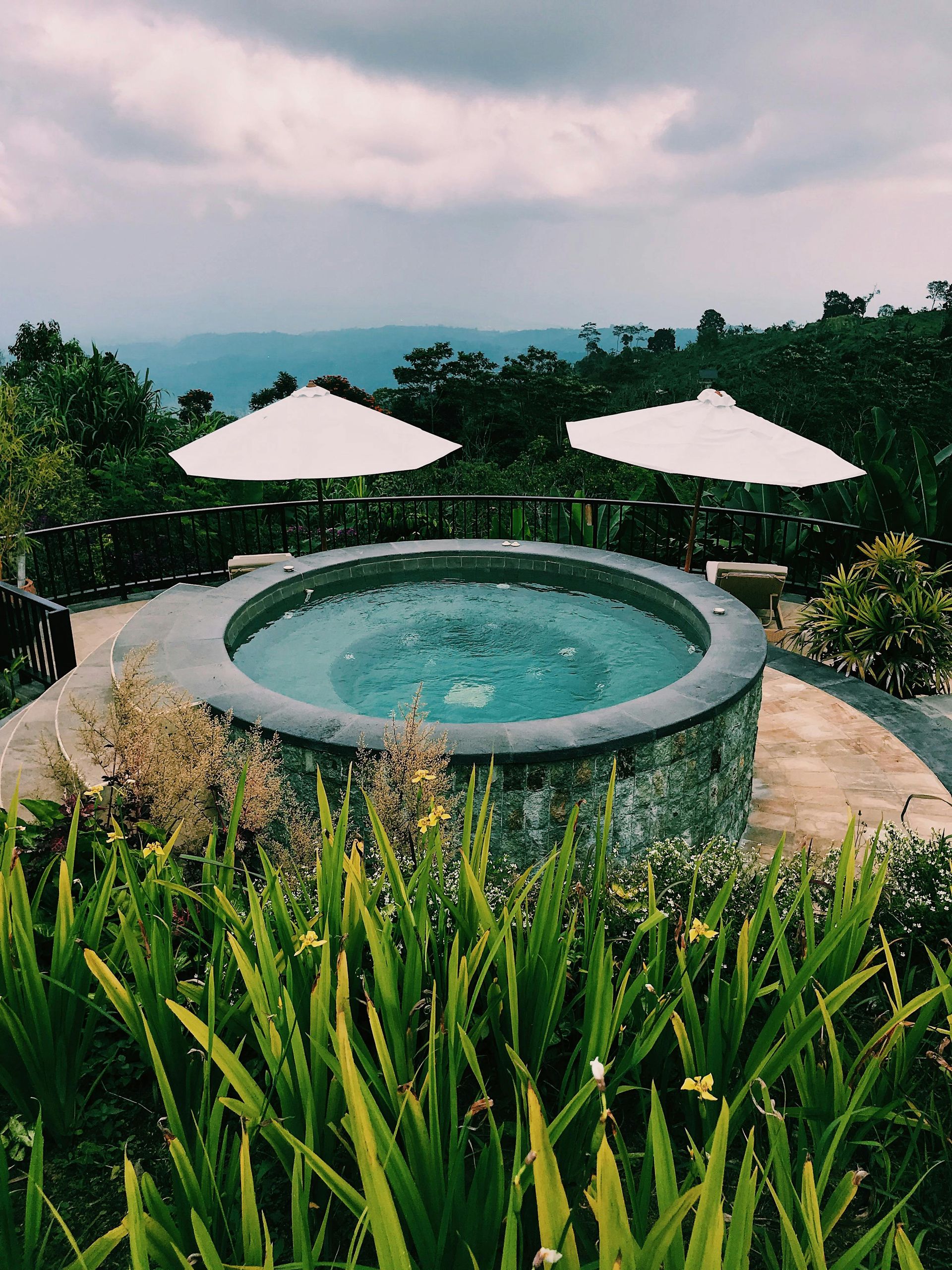 Stone hot tub outdoors with umbrellas, overlooking a landscape.