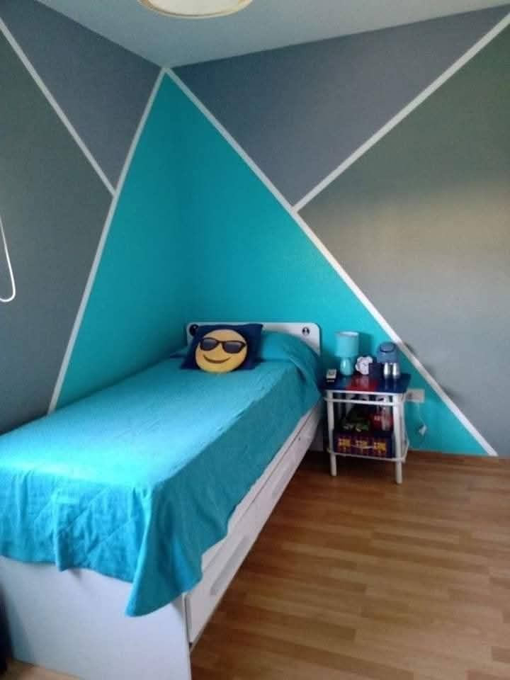 Piccola camera da letto con letto turchese, pareti con motivi geometrici color verde acqua e grigio, e un carrello da comodino con bottiglie.