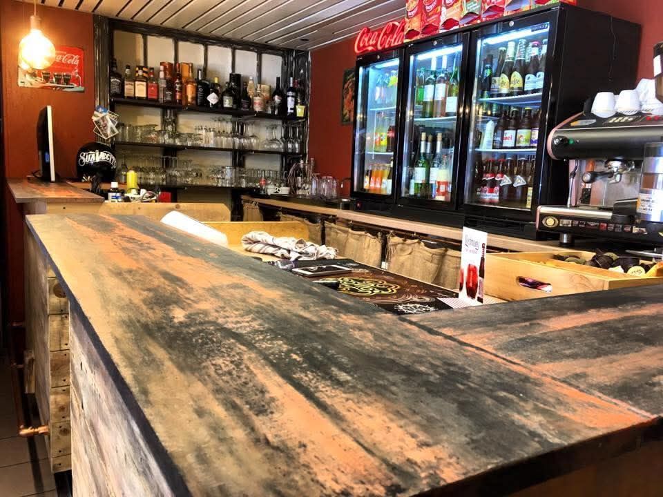Bar rustico con lungo bancone in legno, scaffali per liquori e frigoriferi per bevande illuminati sullo sfondo.