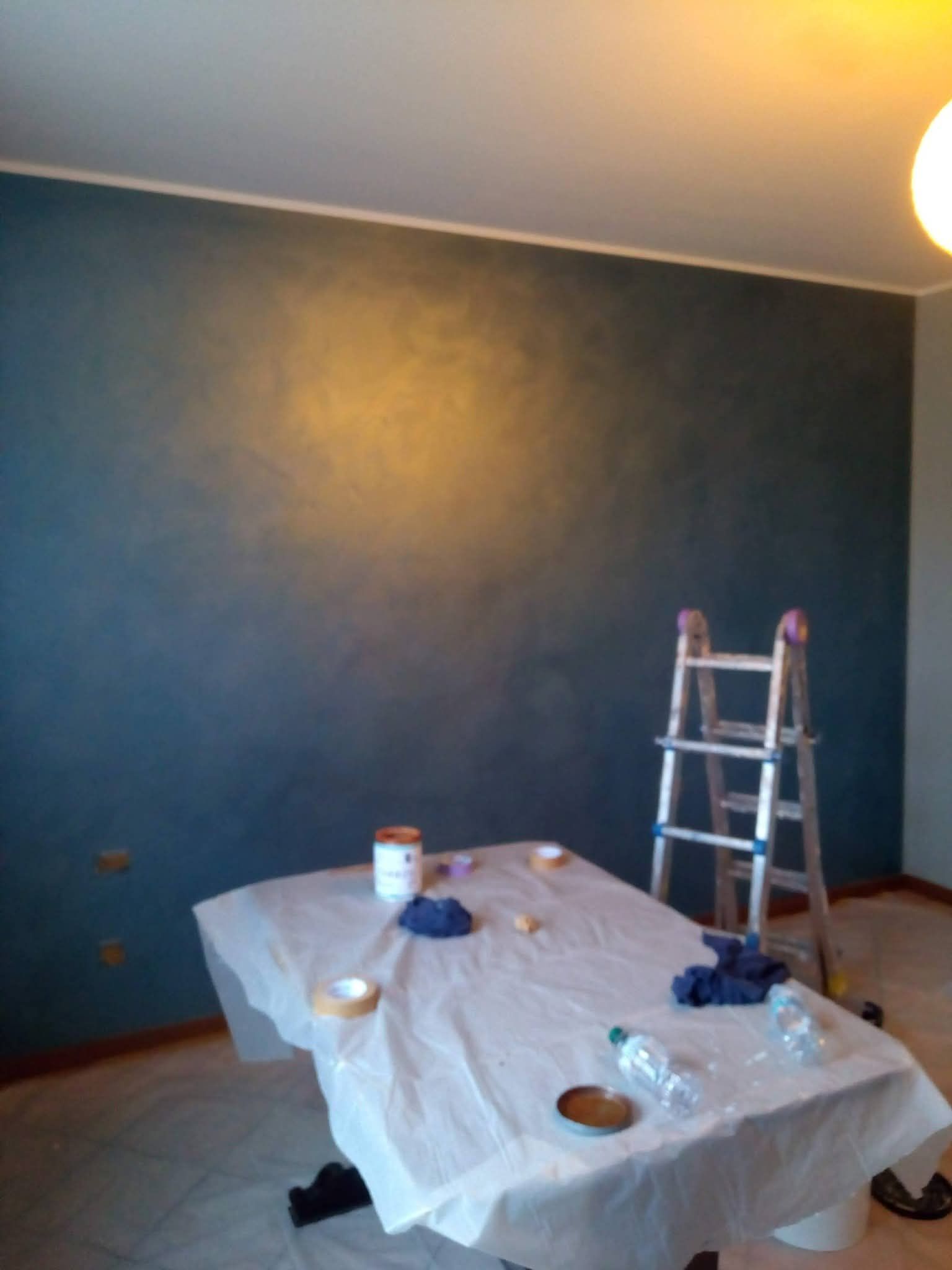 Stanza dipinta di blu scuro con un tavolo coperto da teli protettivi e una scala appoggiata al muro.