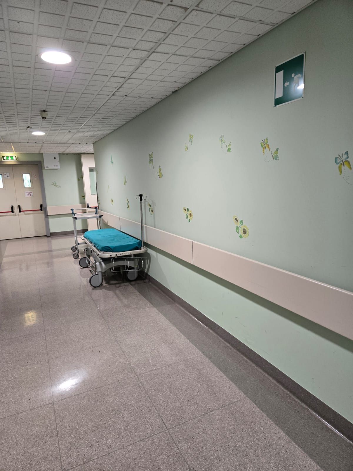 Corridoio ospedaliero vuoto con una barella blu lungo la parete e porte in fondo.