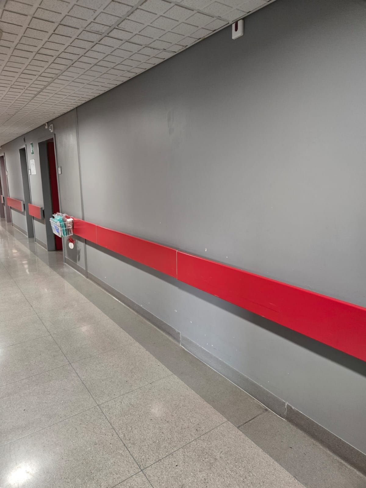 Corridoio ospedaliero vuoto con pareti grigie, corrimano rosso e pavimento piastrellato.