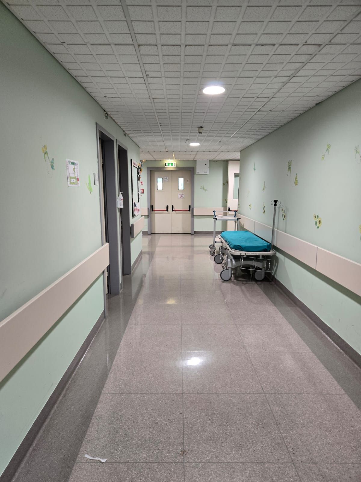 Corridoio dell'ospedale con pareti chiare, luci a soffitto e una barella su ruote in fondo.