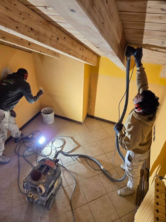 Due operai dipingono un soffitto inclinato in una piccola stanza, con secchi di vernice e cavi sparsi sul pavimento.