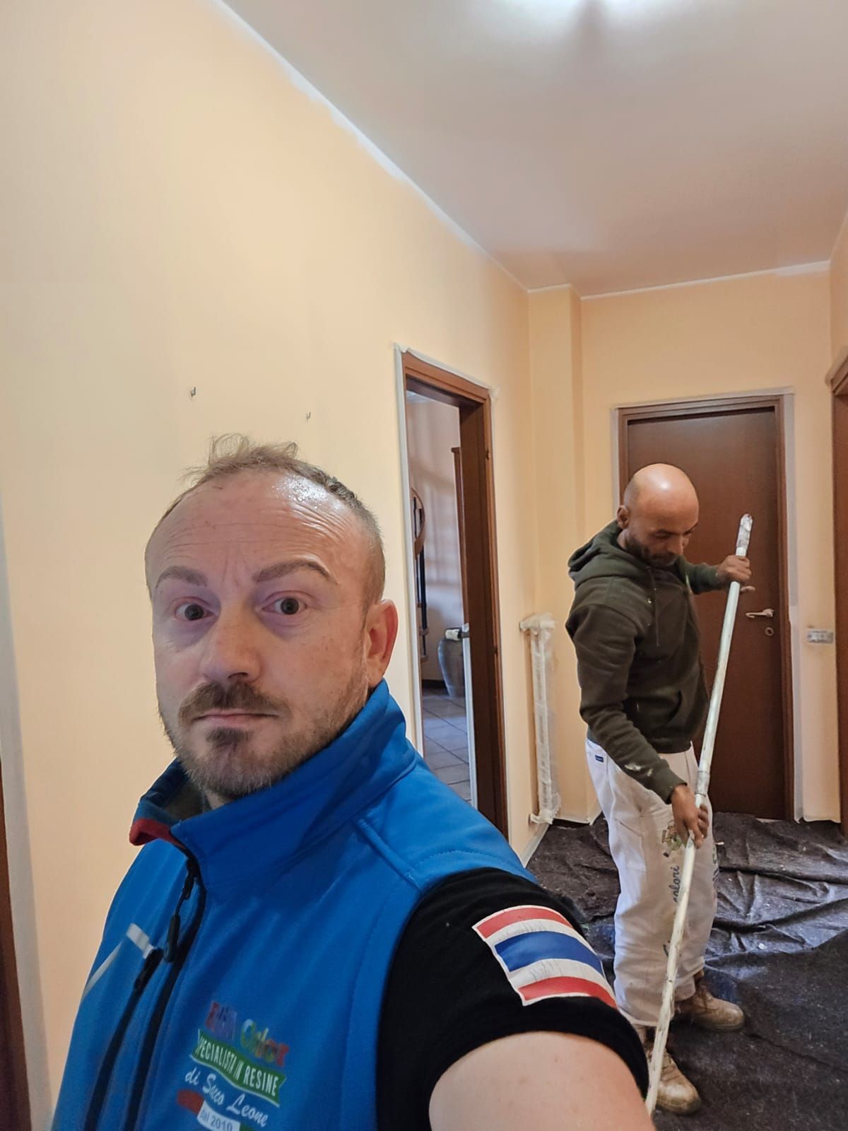 Due operai in un corridoio, uno con un gilet blu in primo piano e l'altro intento a dipingere una porta sullo sfondo.