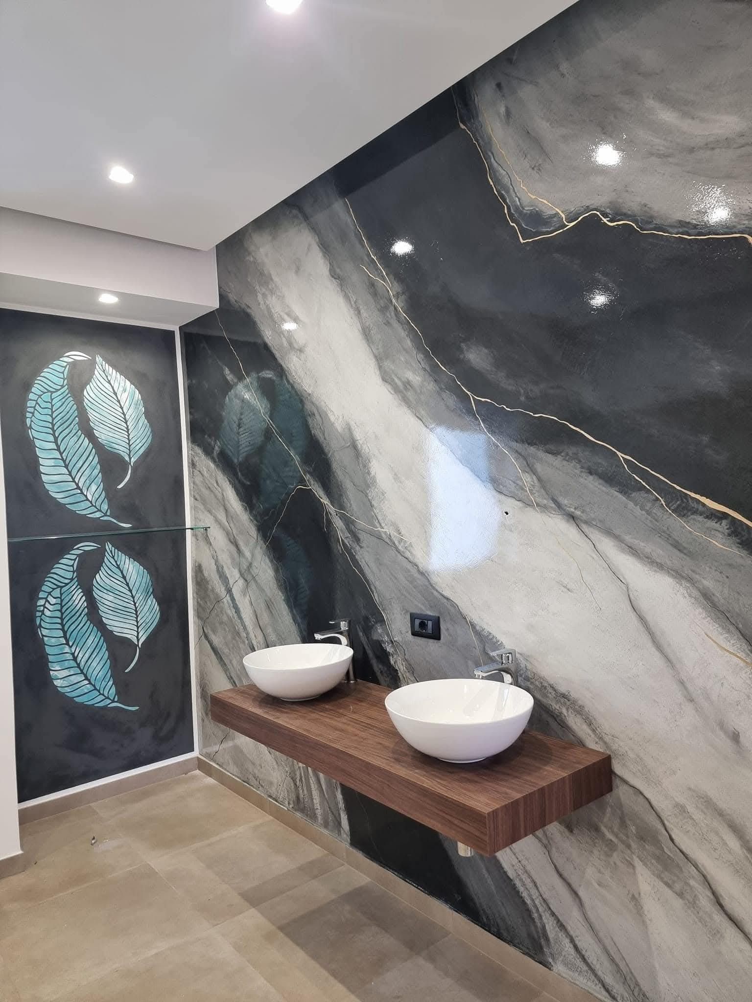 Bagno moderno con parete in marmo, ripiano in legno per il lavabo, due lavabi a ciotola bianchi e pannelli decorativi con motivi a foglie.