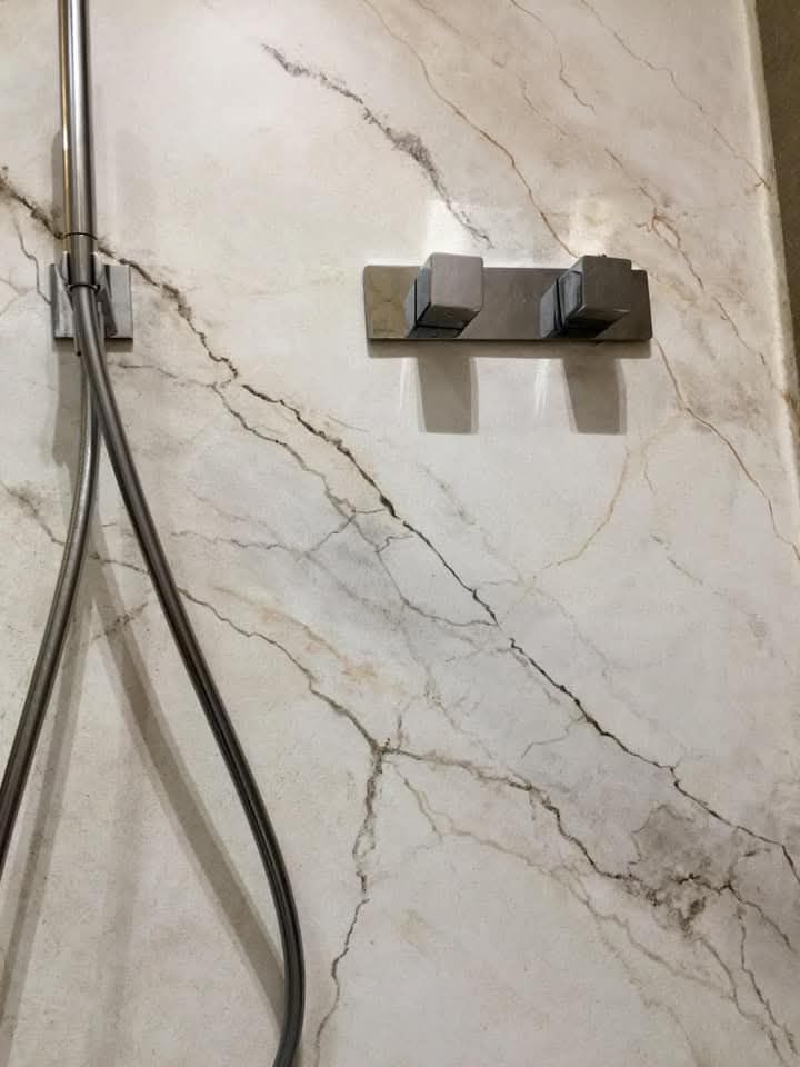 Parete della doccia con motivo effetto marmo, dotata di due ganci metallici e tubi flessibili a sospensione sulla sinistra.
