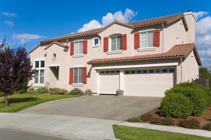 Suburban two-story beige house with red shutters, three-car garage, and a driveway under a blue sky