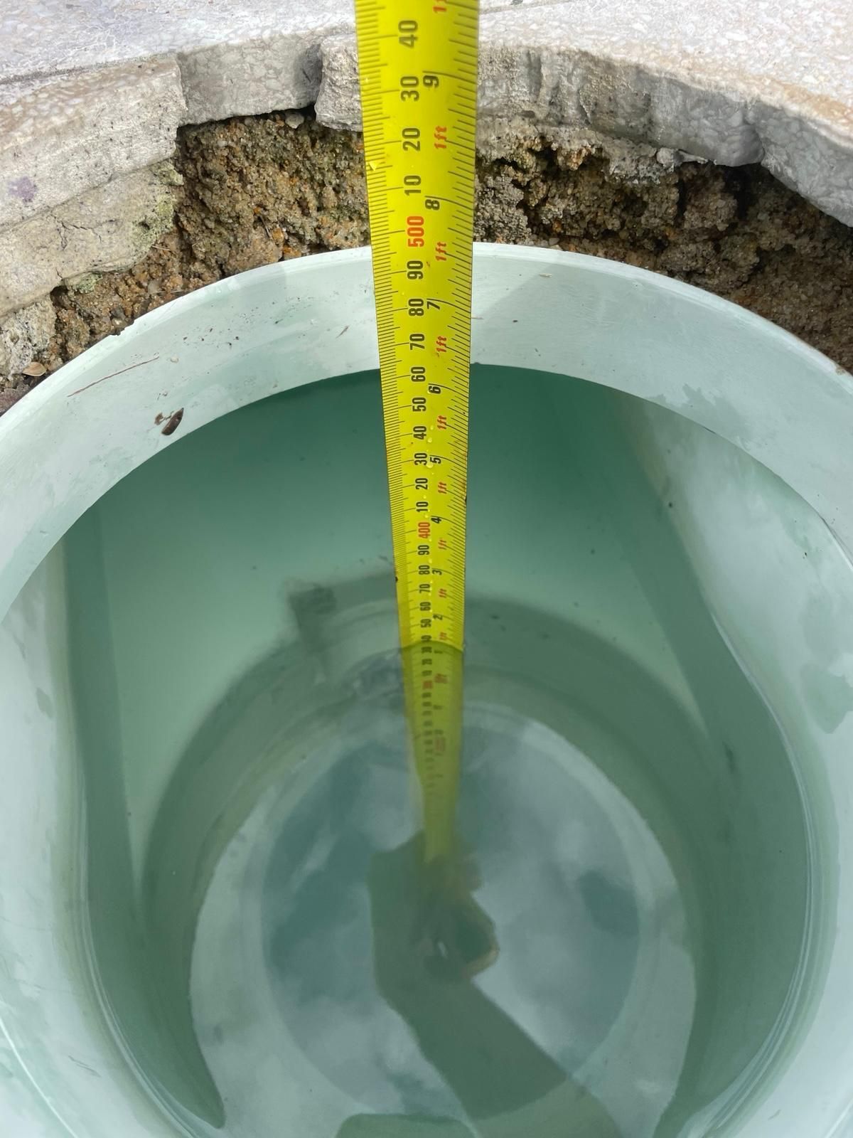A tape measure lowered into a water-filled, light green circular utility pipe set in the ground.