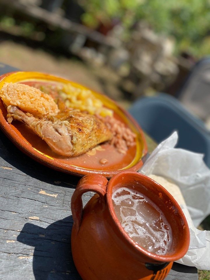 Plate of chicken, beans, potatoes, and a cup of drink, set on a wooden surface outdoors.
